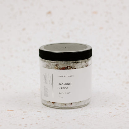 Clear jar holding bath salt mixture with salts and flower petals, with black lid and white label saying "Jasmine and Rose"