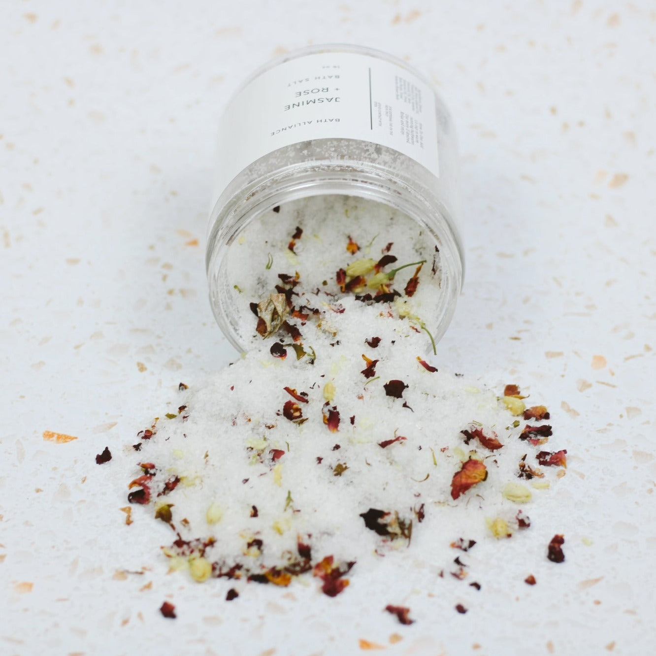 Bath salt jar turned on side, with bath salt mixture of white salts and pieces of flower petals pouring out.