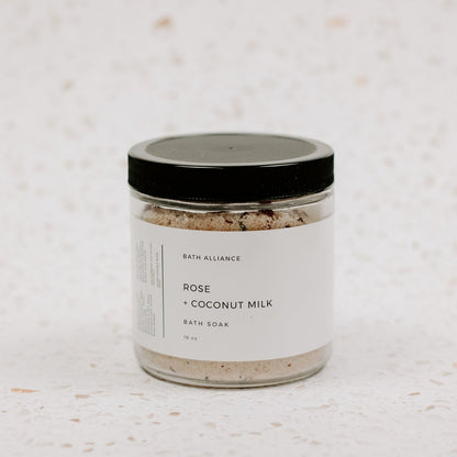 clear jar with dark lid and white label saying "Rose + Coconut Milk bath soak" in plain lettering. Bath soak mixture is inside.