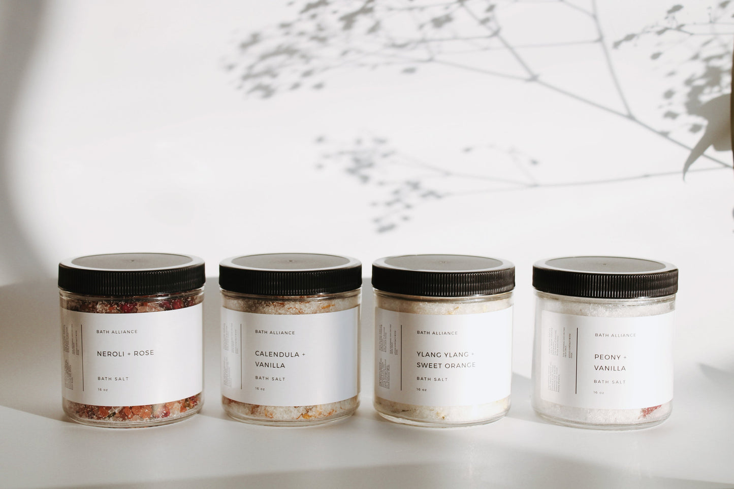 Four clear jars with dark lids, different bath salt mixtures in each of varying color. Each jar has a white label with the title written on it, "Neroli + Rose", "Calendula + Vanilla", "Ylang Ylang + Sweet Orange", and "Peony + Vanilla".
